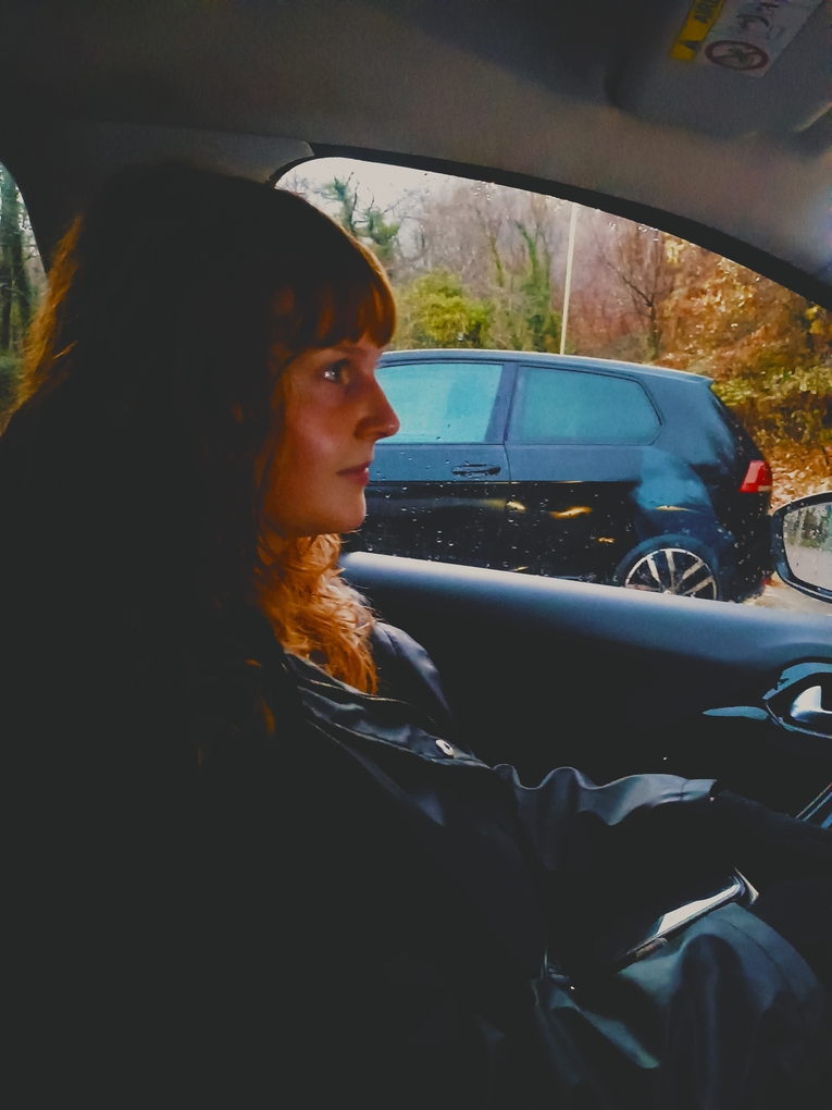Deeply lit photo of a young woman with red hair, she is sat in a car with her face in profile